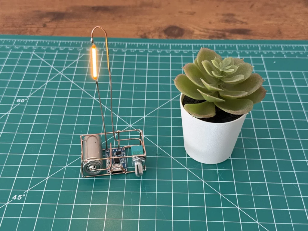 The finished copper wire lamp lit up on the cutting mat, its tall copper arm holding the glowing LED filament beside a potted succulent