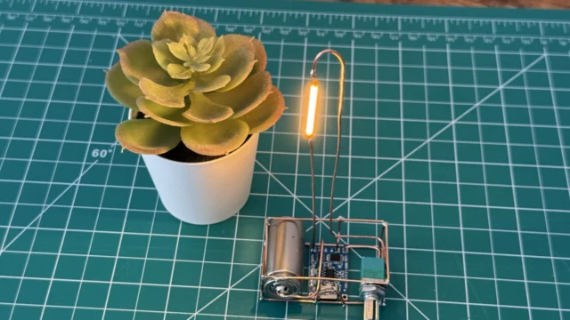 Copper wire lamp glowing warm orange on a green cutting mat next to a small succulent, with the battery, charger board and dimmer knob in its copper base