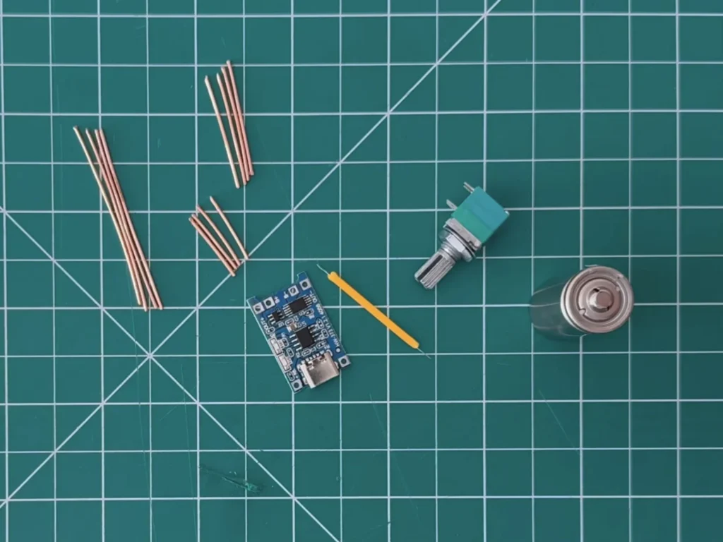 Lamp parts on a green cutting mat: straight copper rods, a TP4056 USB-C charger board, a yellow LED filament, a rotary potentiometer and a 16340 battery