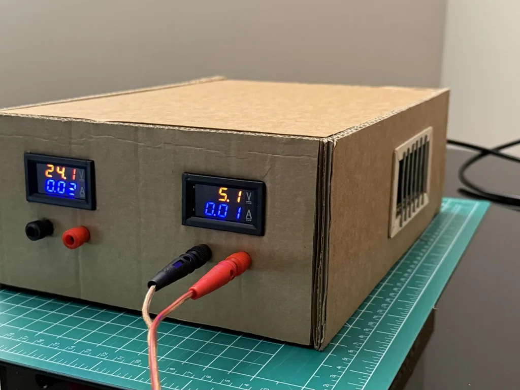 Front of the cardboard bench power supply with both meters lit and test leads plugged into the 5 V output