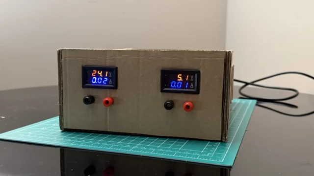 Finished DIY bench power supply in a cardboard case, showing 24.1 V and 5.1 V on its two digital meters with test leads plugged into the banana terminals