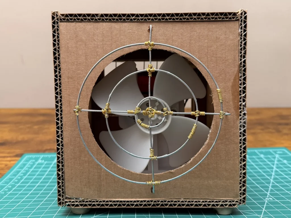 Front of the finished DIY desk fan: a cardboard box with a round opening, the white fan blade behind a stainless steel wire guard with gold aluminium wraps