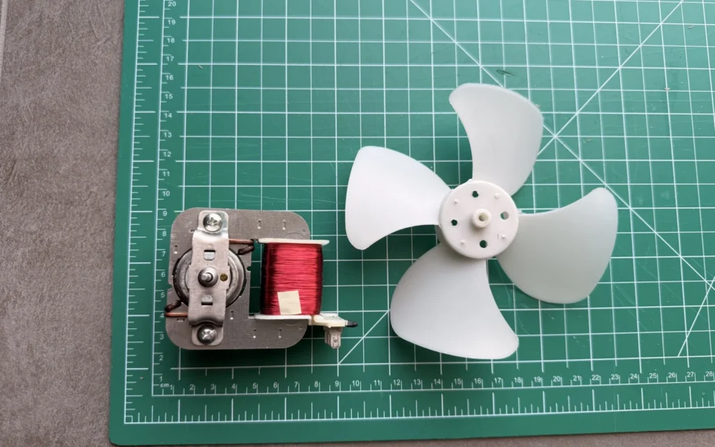 Salvaged microwave fan motor with its red copper coil next to the white four-blade fan on a green cutting mat