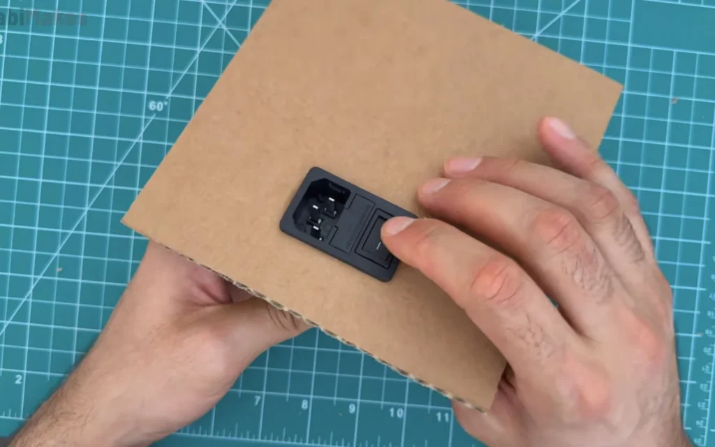 Black power inlet module with fuse holder and rocker switch placed on a cardboard panel to mark its cut-out