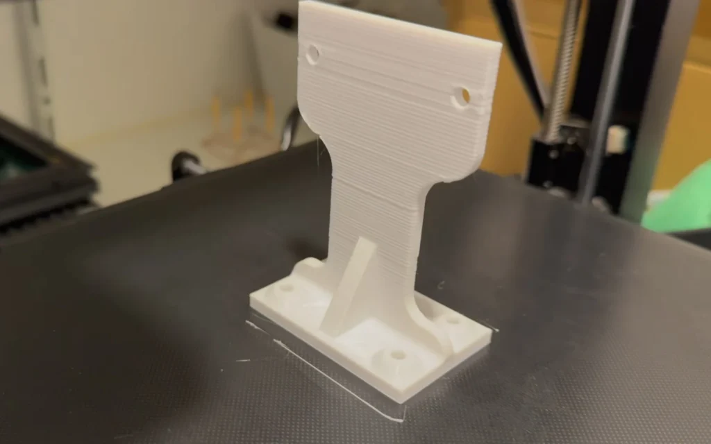 The first 3D-printed motor mount on the printer bed: a thin upright plate with two holes, a narrow neck and a foot with four screw holes