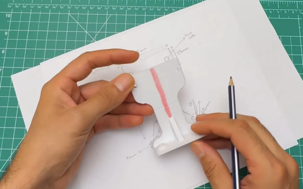 Hand holding the first printed motor mount over the paper sketch, with the weak neck area marked in pink