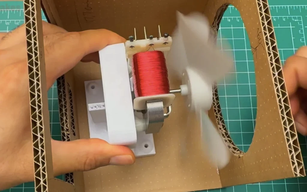 Hand holding the fan motor on its printed mount inside the cardboard box, with the blade spinning behind the round opening