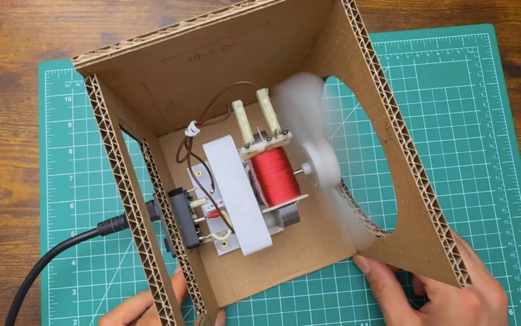 Top view into the cardboard box: the motor on its printed mount, the wired inlet module in the back panel and the blade spinning in the front opening