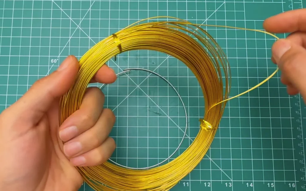 Hands unwinding gold-coloured aluminium craft wire next to a stainless steel wire ring
