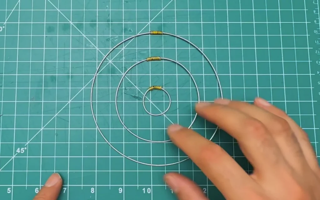 Three concentric stainless steel wire rings on a cutting mat, each joined with a wrap of gold aluminium wire