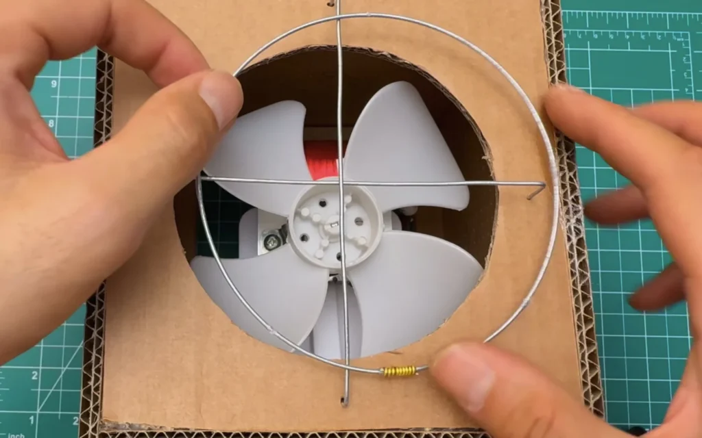 Stainless steel ring with a wire cross held over the fan opening, the hooked ends going into small holes in the cardboard