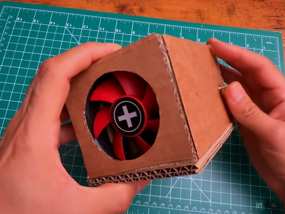 Finished cardboard fume extractor held in two hands, red fan visible through the round front opening and the toggle switch on the side