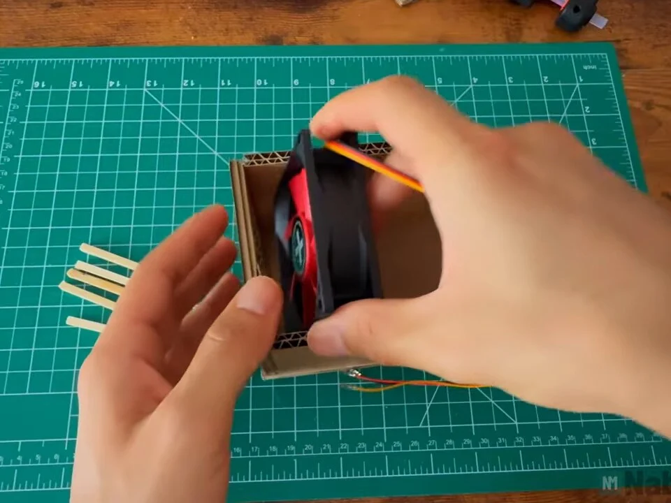 PC fan being slid into the cardboard box behind the front opening, with cut sticks on the mat