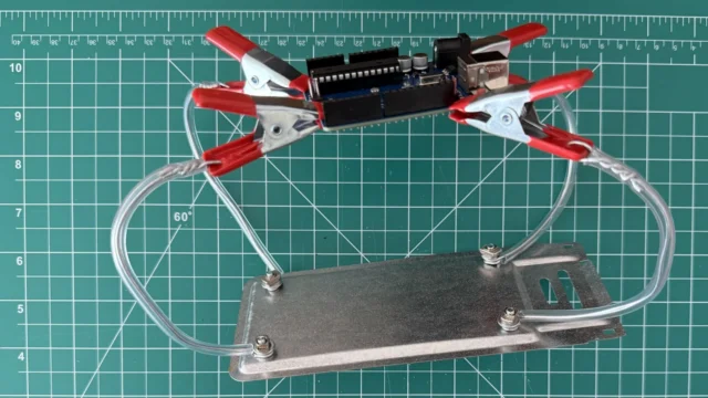 DIY helping hands holding an Arduino Uno with four red spring clips on bendable wire arms bolted to a metal base plate