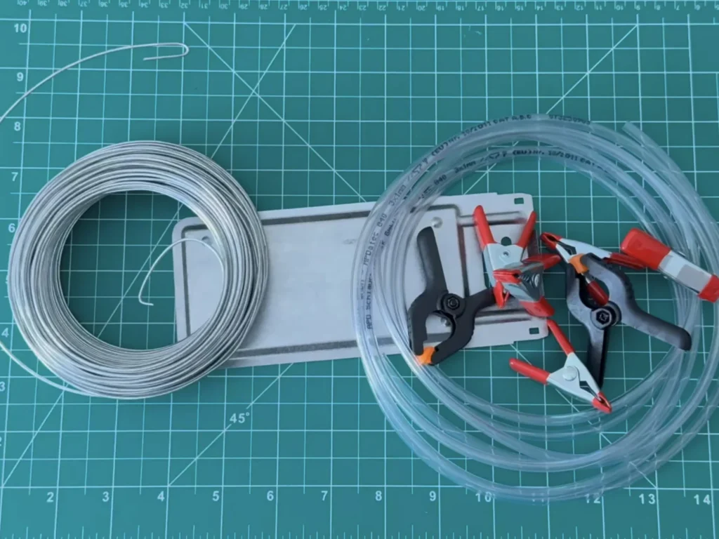 Materials for the DIY helping hands on a cutting mat: a coil of steel wire, a flat metal plate, clear tubing and red spring clips