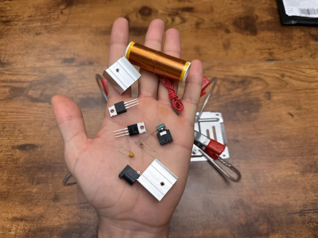 Mini Tesla coil kit parts in an open hand: the copper secondary coil, two aluminium heatsinks, the IRF530 and TIP41, the audio and DC jacks, a capacitor and the neon bulb