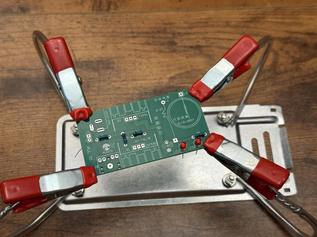 Green Tesla coil PCB held in helping hands with red clips, the 10 kΩ and 2 kΩ resistors and two red LEDs fitted