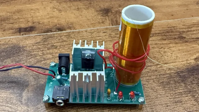 Finished mini Tesla coil on a wooden desk: the orange secondary coil wrapped in the red primary, the transistors on aluminium heatsinks, and the DC and audio jacks