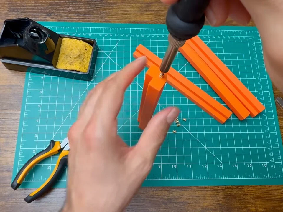 Soldering iron pressing a brass heat-set insert into an orange printed frame corner