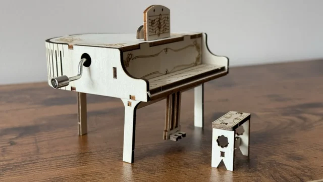 Finished wooden piano music box on a wooden desk, with the hand crank on its side, an engraved music stand and a tiny matching bench