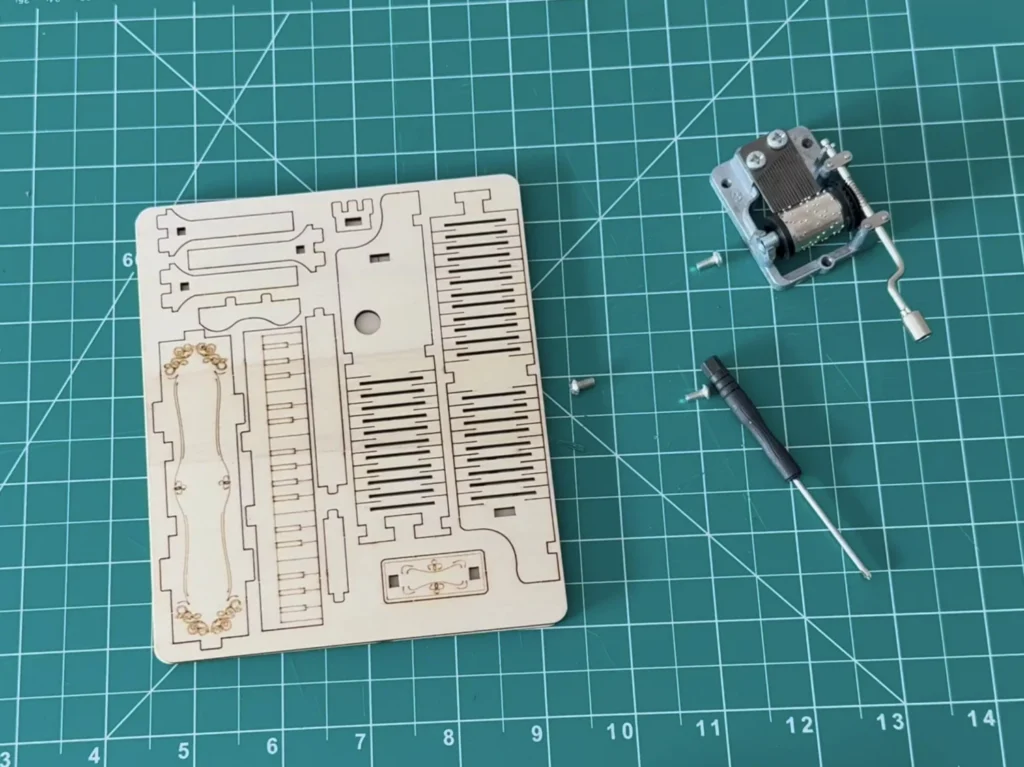 Wooden piano kit laid out on a cutting mat: the laser-cut plywood sheet with engraved parts, the hand-crank music box movement, small screws and a screwdriver