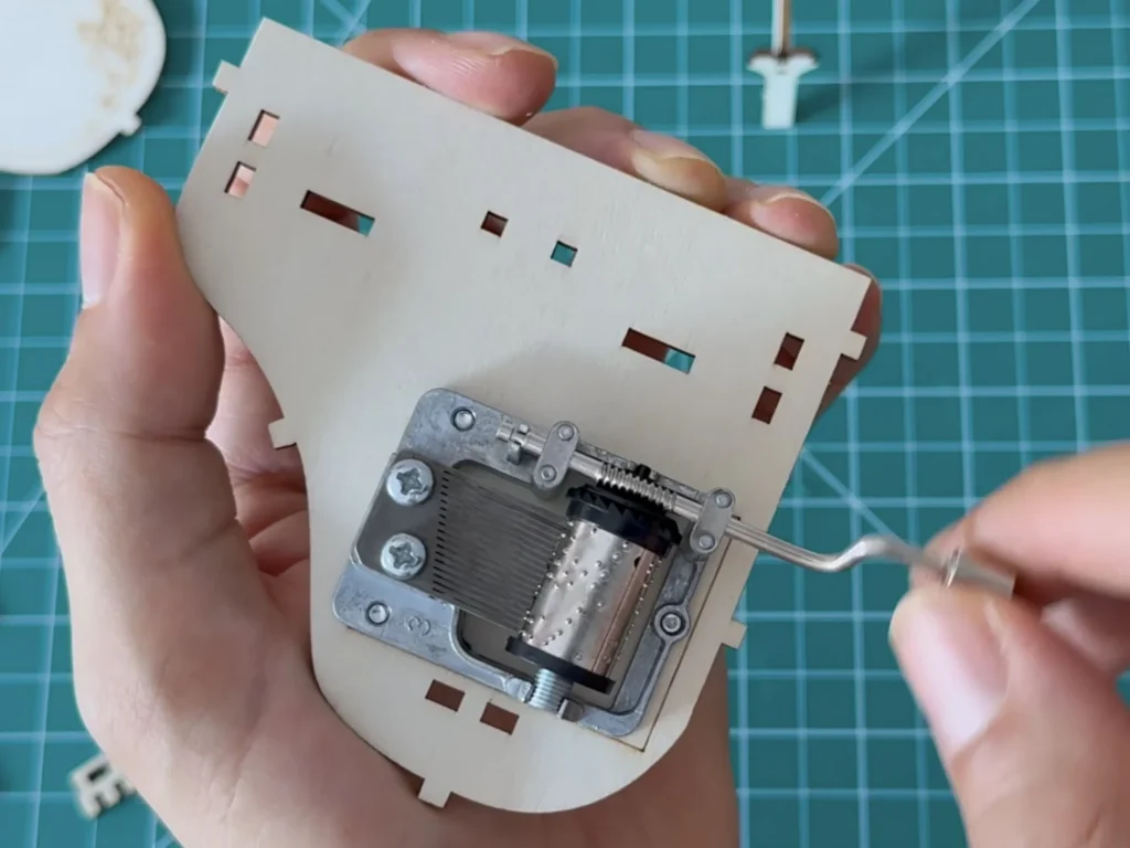Hand holding the curved plywood base plate of the piano with the metal hand-crank music box movement screwed onto it