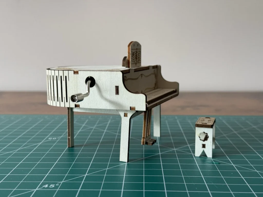 Side view of the finished wooden piano music box at eye level, with the crank handle in the side of the body and the bench beside it